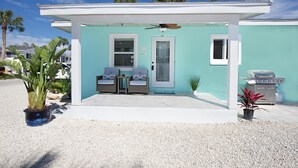 A small, single-story building with a covered patio area, two wicker chairs, a small table, a door, and a grill. - Coconut Villa A (Fort Myers Beach)