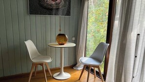 Une pièce avec deux chaises blanches disposées autour d’une petite table ronde sur laquelle se trouve un vase en verre. Le sol est en bois, un tableau est accroché au mur, et une fenêtre offre une vue sur la verdure.