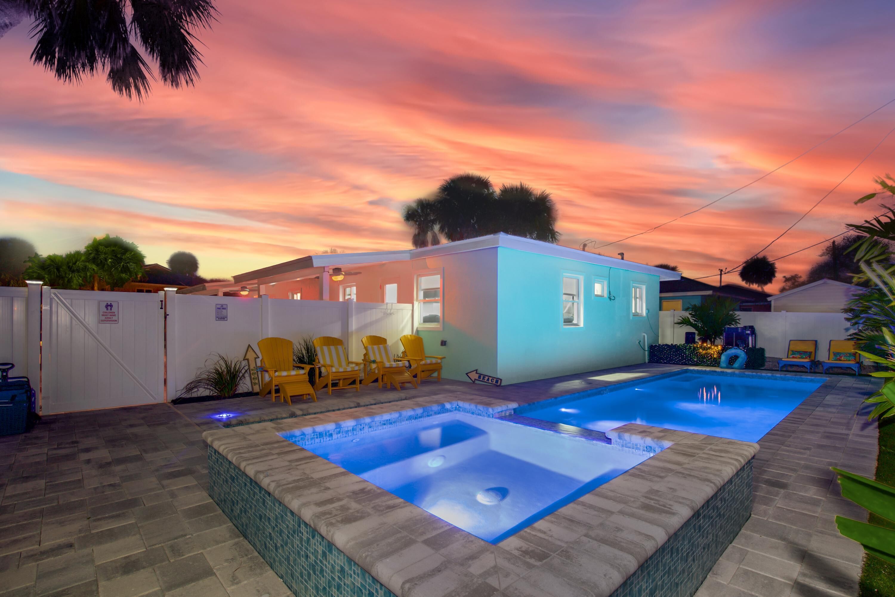 A swimming pool with a hot tub, surrounded by a fenced area with yellow chairs, under a sunset sky.