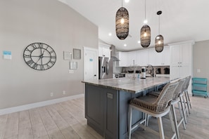 A modern kitchen with a central island, bar stools, pendant lighting, and a large wall clock. - Harborside on Palermo (Fort Myers Beach)