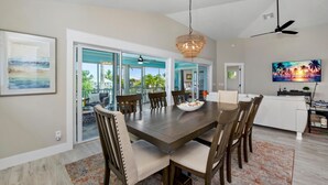A dining area with a large wooden table, six chairs, and a chandelier. There is a television on the wall and a door leading to a balcony with a view of palm trees and a house. - Harborside on Palermo (Fort Myers Beach)