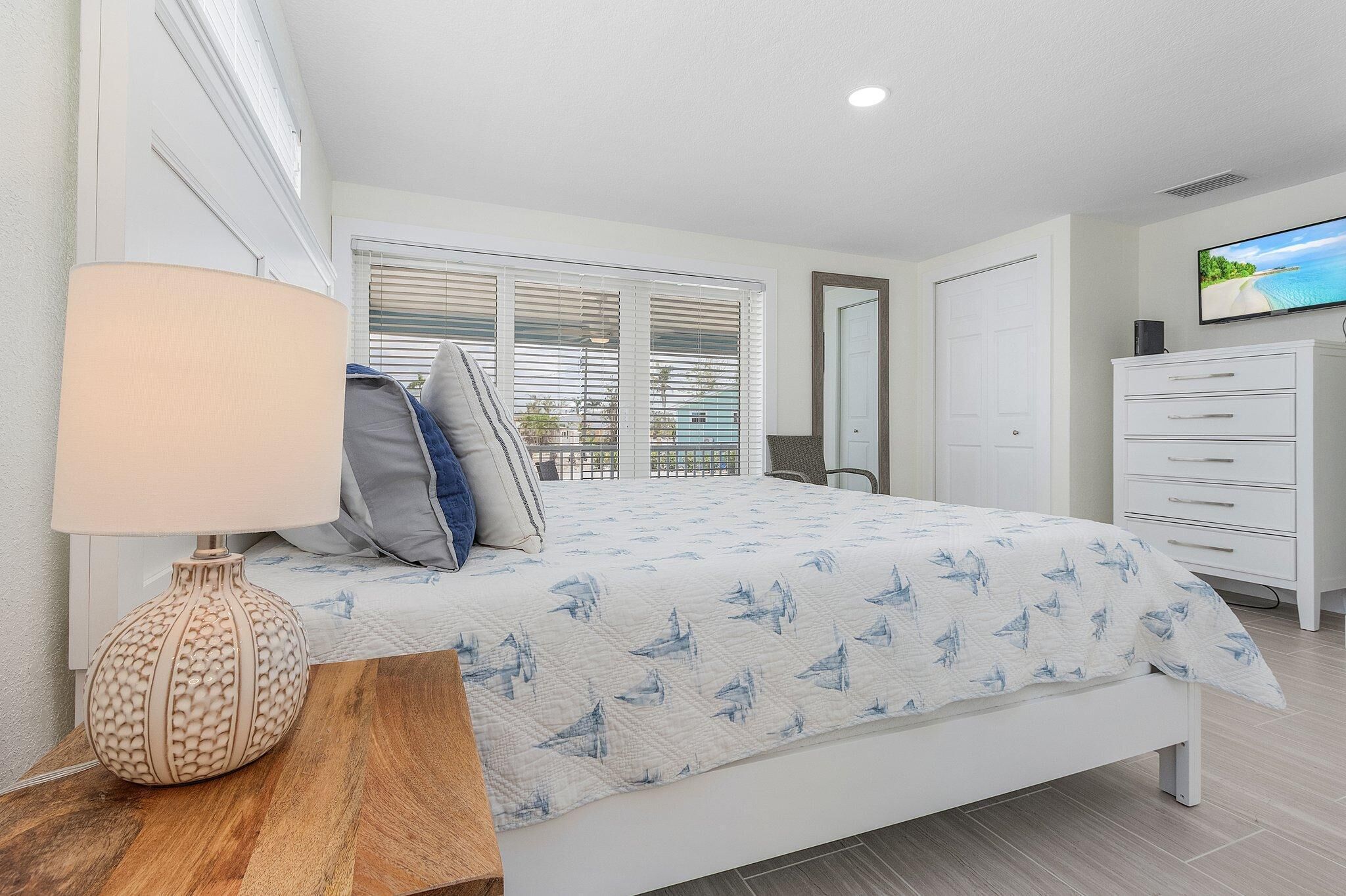 A bedroom with a bed, a bedside table with a lamp, a dresser, and a TV on the wall.