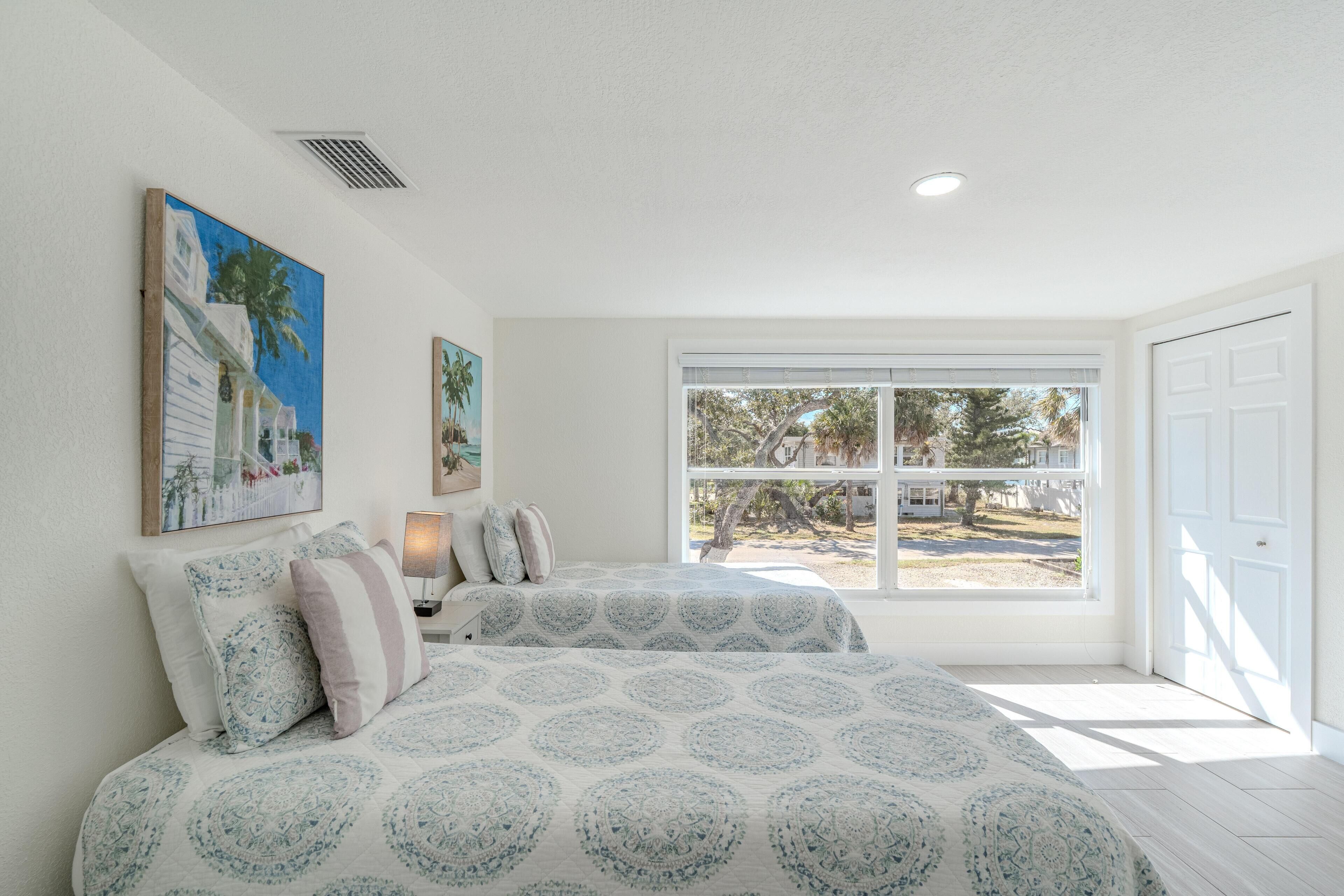 A bedroom with two beds, a large window, and artwork on the walls.