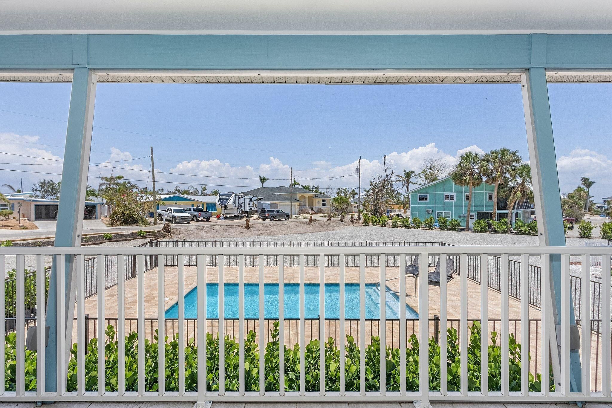 A view of a swimming pool area with a wooden deck, surrounded by a white fence and green plants.