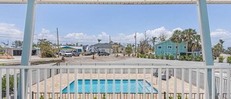 A view of a swimming pool area with a wooden deck, surrounded by a white fence and green plants.