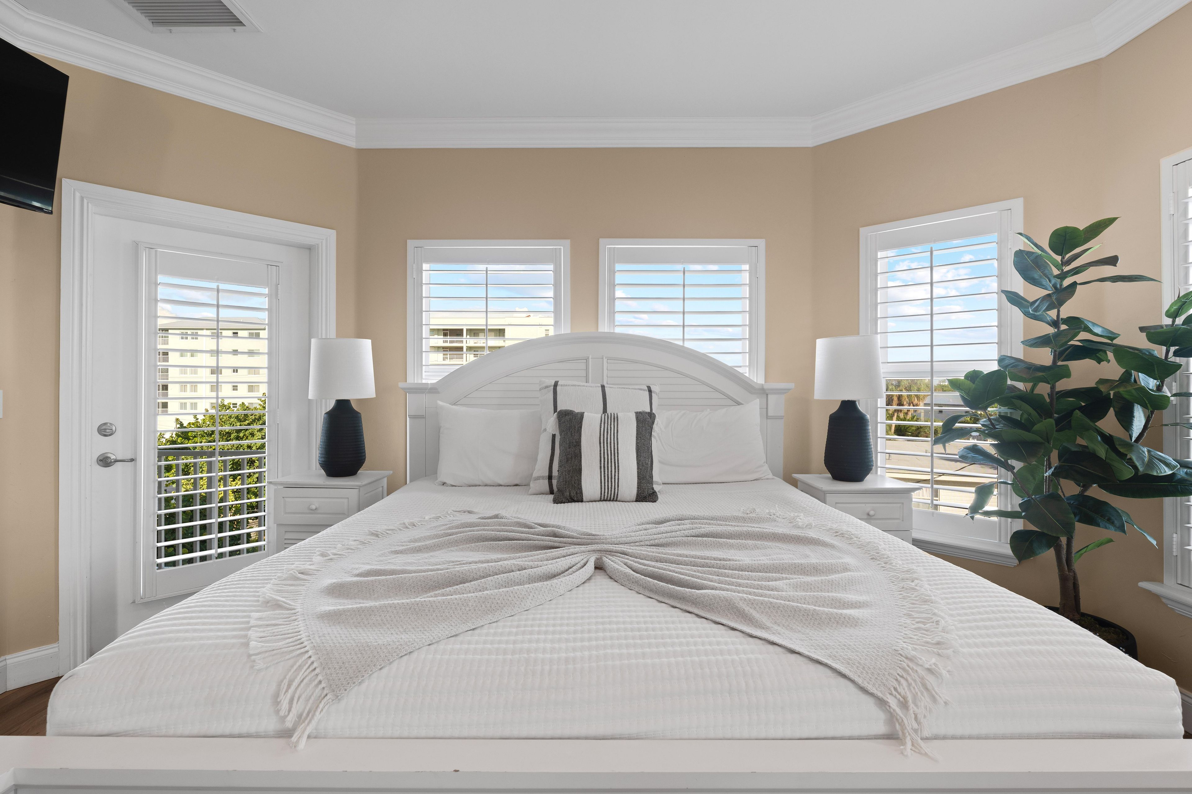 A bedroom with a large bed, two bedside tables, two lamps, a window with blinds, and a potted plant.