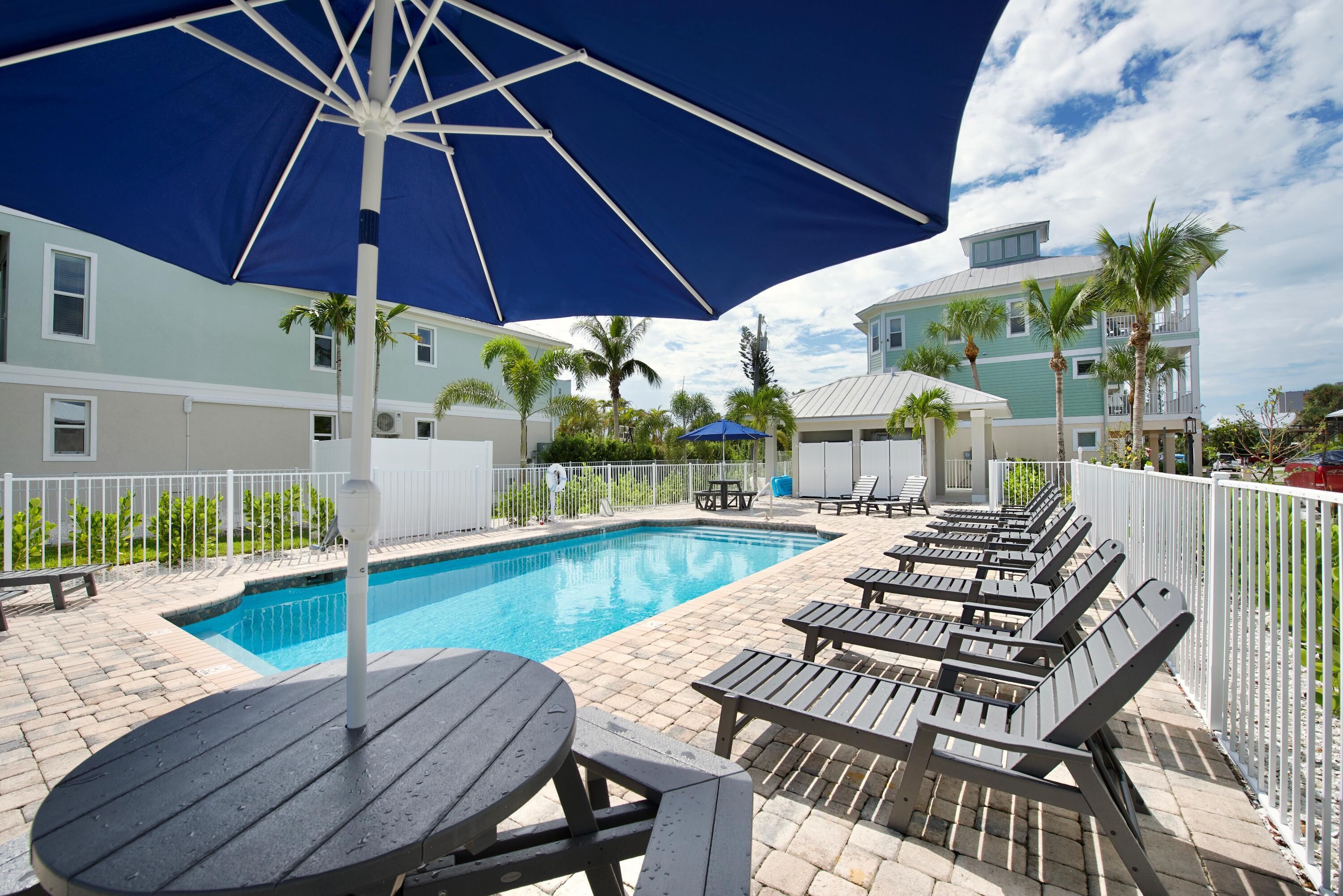 A swimming pool with lounge chairs and umbrellas, surrounded by a white fence and palm trees.
