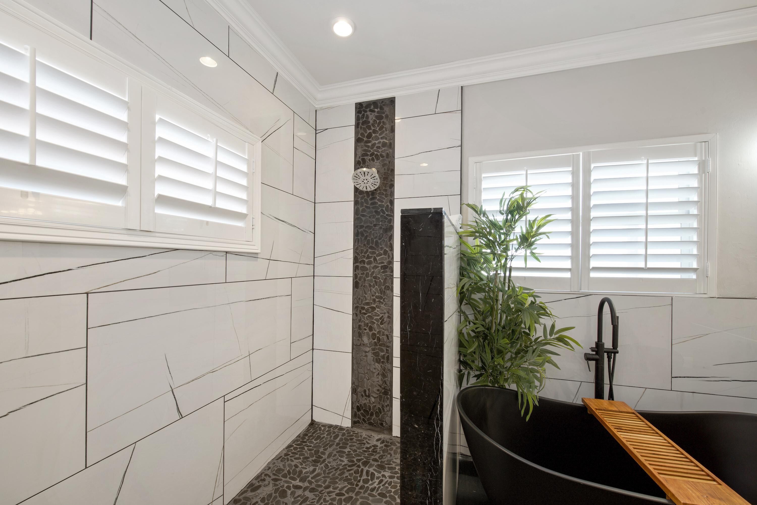 Un baño con paredes de azulejos blancos, una bañera negra, un banco de madera, una planta en maceta y una ventana con persianas.