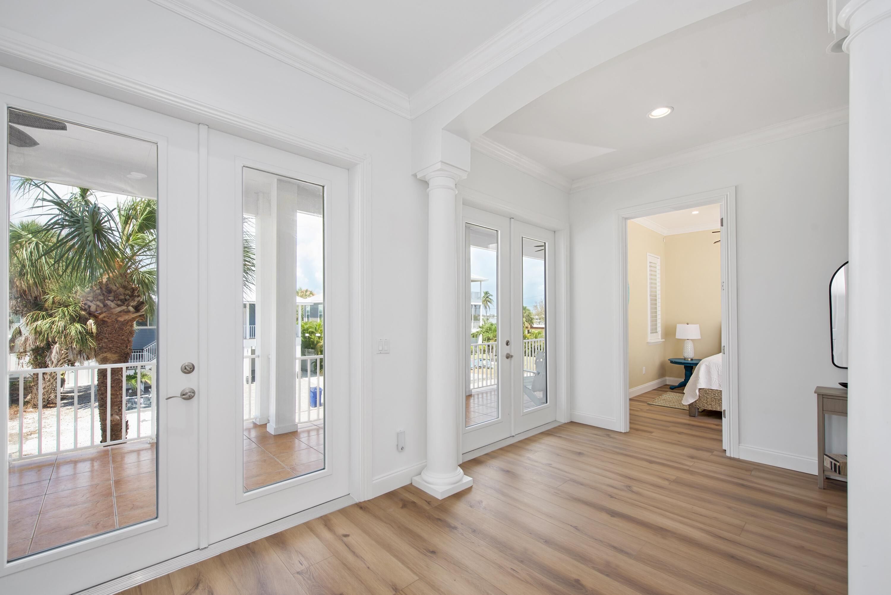 A spacious hallway with wooden flooring, white walls, and large glass doors leading to an outdoor area with a palm tree.