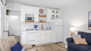 Cocina funcional con armarios blancos, microondas, estufa y refrigerador. Hay un tostador azul en la encimera y una pequeña mesita de noche con lámpara.