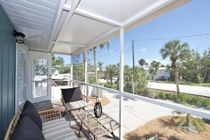 Relax in a serene outdoor space with wicker furniture, a glass table, and a lantern-style light fixture, surrounded by palm trees and a residential view.