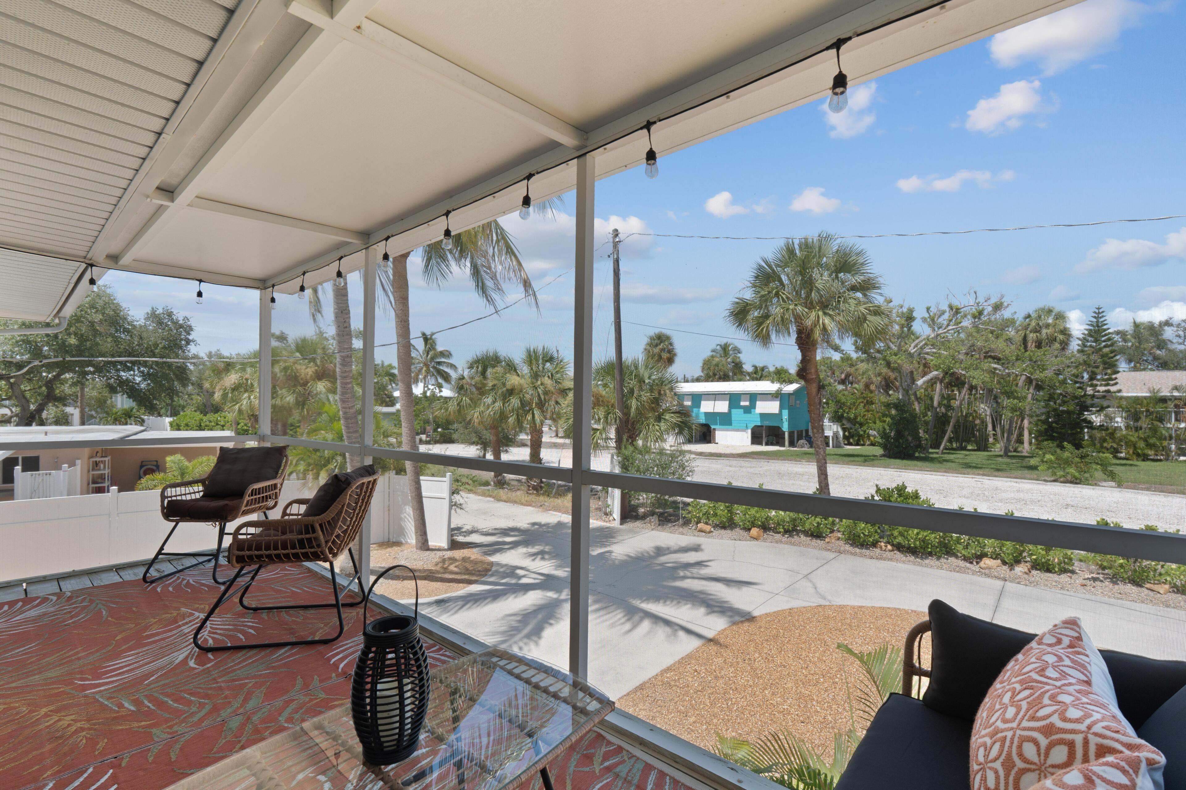 A covered outdoor seating area with chairs and a table, overlooking a street with palm trees and a building.