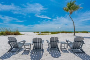 Een strand met vier Adirondack stoelen en een tafel, een palmboom, en een heldere blauwe lucht.