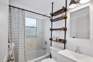 Een moderne badkamer met bad, toilet en wastafel. Er is een raam met uitzicht op de lucht, en een douchegordijn met een bladmotief. Boven het toilet zijn planken bevestigd aan de muur.