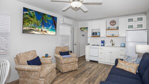 A modern living room with a ceiling fan, a large beach-themed painting, a kitchen with white cabinets, and a blue sofa. - Lucky Charm 2 (Fort Myers Beach)