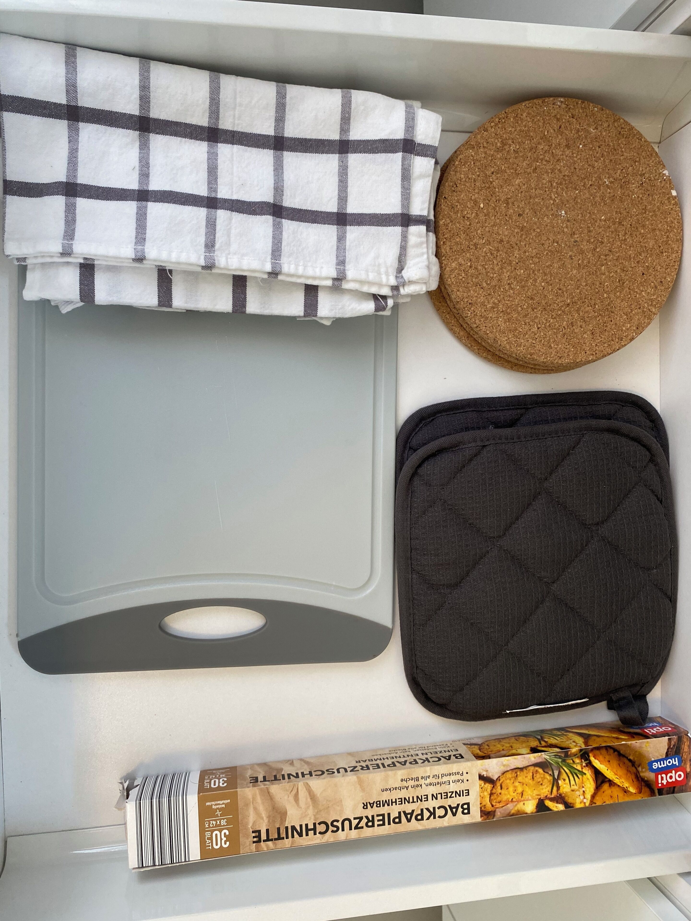 Eine Küchenschublade mit einer grauen Ablage, einer schwarzen gesteppten Tasche, einer runden Korkmatte, einem schwarz-weißen karierten Tuch und einer Packung Gebäck.