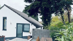 A white building with a dark roof, a door, and a window. There is a paved area with a wooden table and chairs, and a potted plant. - Modern vacation home in the countryside and close to the city! (Koblenz)