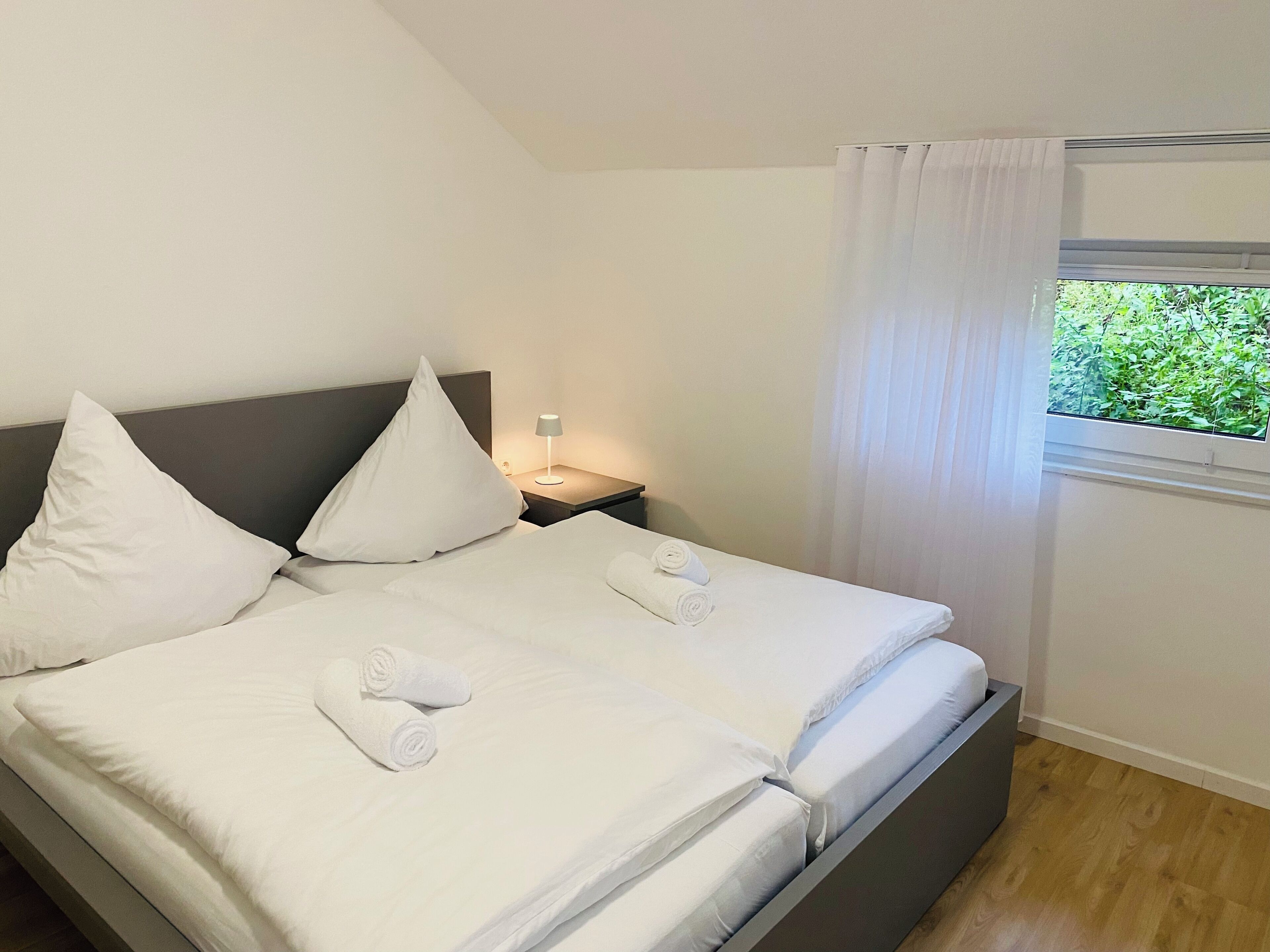 Ein Doppelbett mit weißen Laken und Kissen, ein Nachttisch mit Lampe und ein Fenster mit Vorhängen.