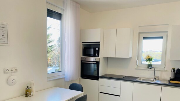 A modern kitchen with white cabinets, a black oven, a microwave, and a sink. There is a window with a view of greenery outside. - Modern vacation home in the countryside and close to the city! (Koblenz)