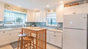 A modern kitchen with white cabinets, a wooden bar stool, a refrigerator, and a window with a view. - Lone Palm Retreat (Fort Myers Beach)