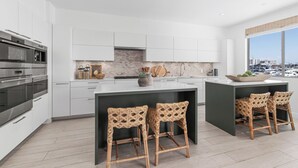 A modern kitchen with white cabinetry, a central island with bar stools, and a view of the marina through large windows. - The Kelly Residence @ Peninsula Village (Newport Beach)