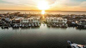 A marina with multiple boats docked, surrounded by buildings and palm trees, with the sun setting in the background. - The Kelly Residence @ Peninsula Village (Newport Beach)