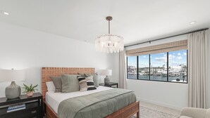 A modern bedroom with a large bed, a wooden headboard, a chandelier, a nightstand with a lamp, a potted plant, and a view of a marina through a large window. - The Kelly Residence @ Peninsula Village (Newport Beach)