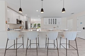 A modern kitchen with white cabinetry, a central island with bar stools, pendant lighting, and a view of the outdoors through large windows.
