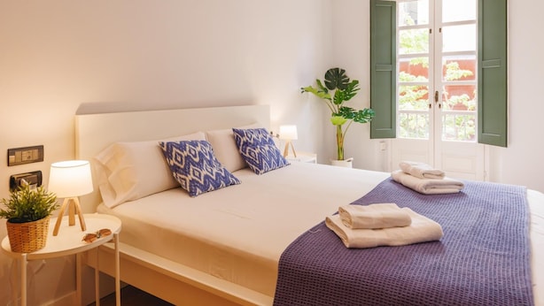 Un dormitorio ordenado con cama, mesitas de noche, lámpara y ventana con contraventanas.