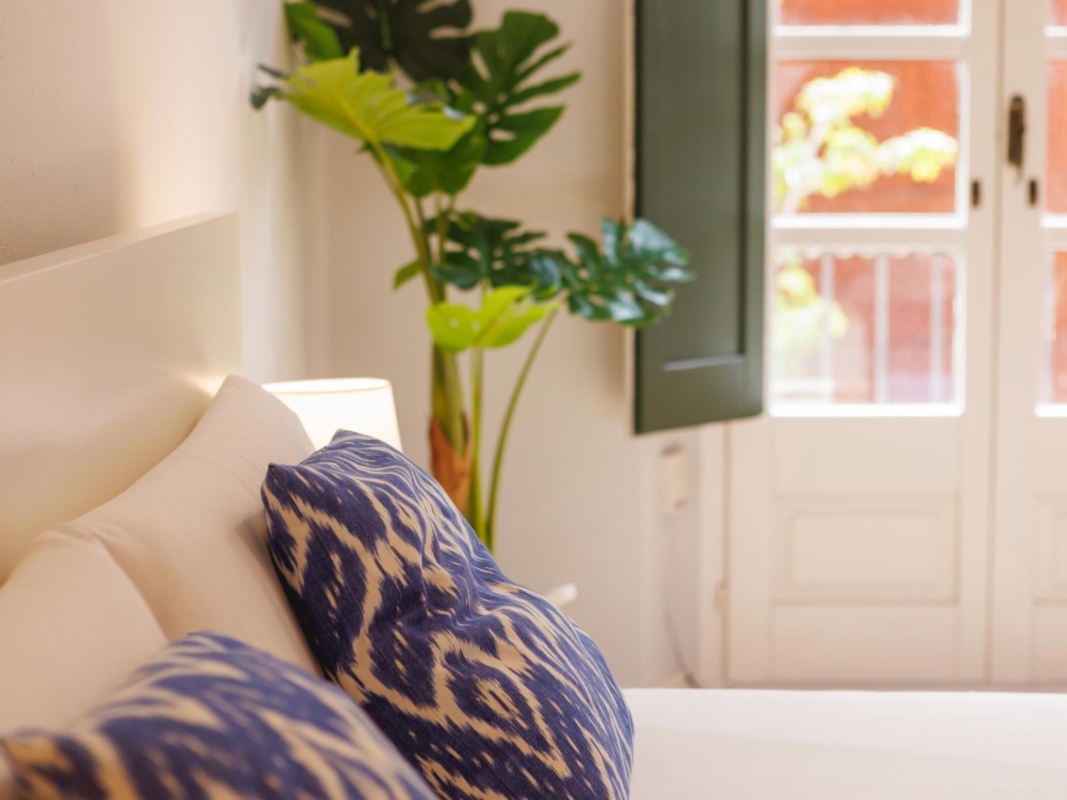 A bed with white and blue patterned pillows, a bedside plant, and a view of a white door with a window.