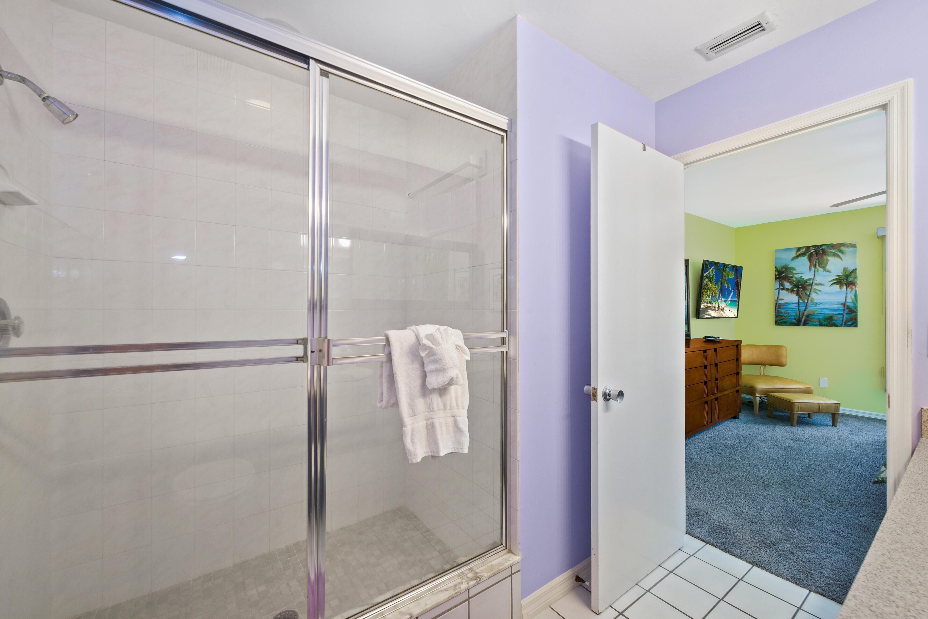 A modern bathroom with a glass shower enclosure, a white towel hanging on the door, and a view into a living area with a blue carpet and a painting of palm trees.