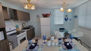 A modern kitchen with wooden cabinets, a stainless steel range hood, and a built-in oven. There is a dining area with a glass table and white chairs, and a window with blinds. - Villa Santorini 3-bedroom apartment in welcoming Bayamón with WiFi, AC. Enjoy  (Bayamón)