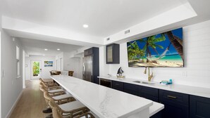 A modern kitchen with a long island, dark cabinets, and a large painting of a beach scene. - Gone Coastal (Fort Myers Beach)