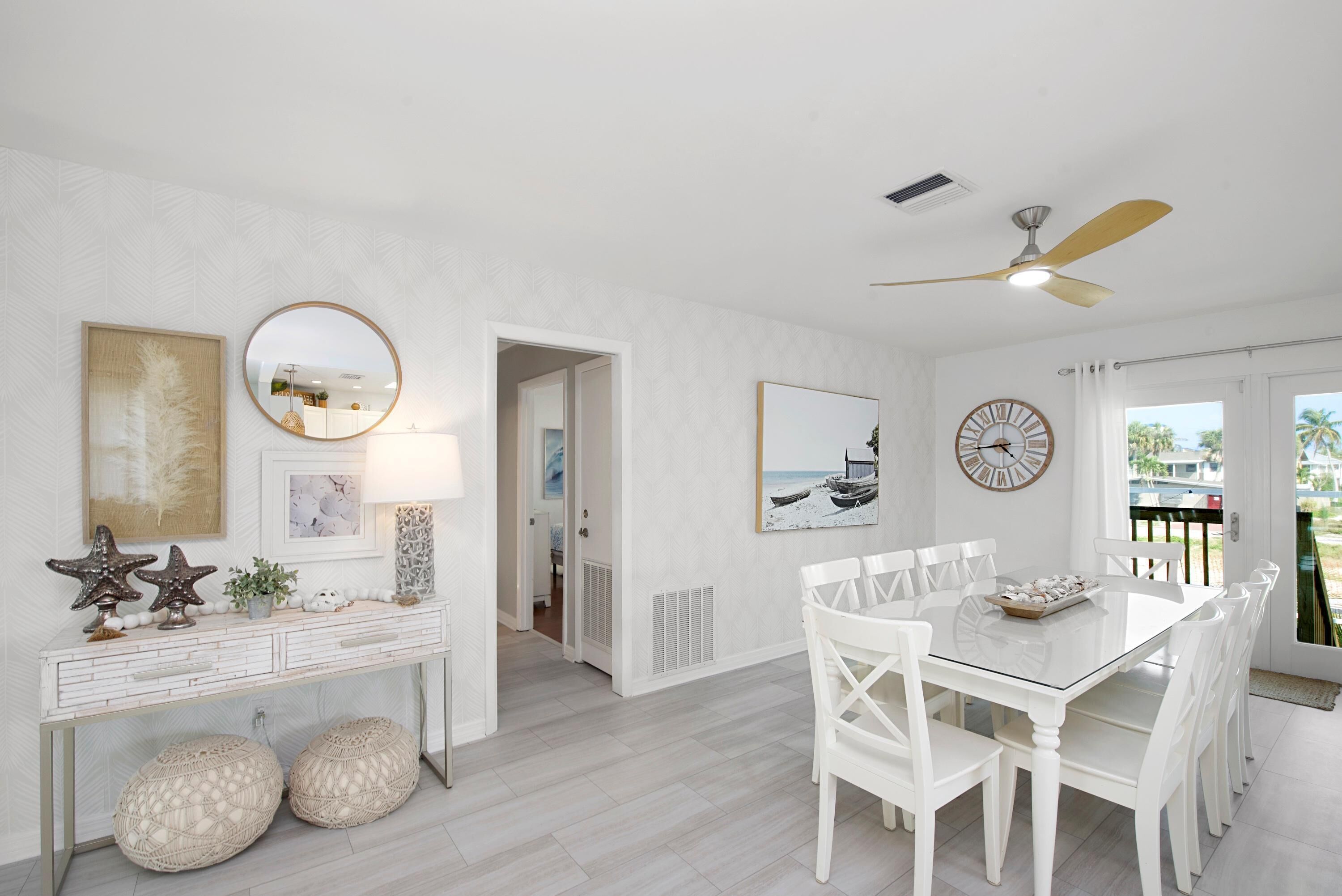 A bright, white interior with a dining area featuring a glass table and white chairs, a ceiling fan, and artwork on the walls.