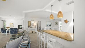 A modern living room with a striped sofa, white chairs, and a kitchen area with a granite countertop. - Gone Coastal (Fort Myers Beach)