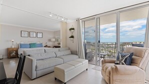 A modern living room with a sofa, ottoman, and armchair, a dining table, and a balcony with a city view. - Estero Beach & Tennis 901-c (Fort Myers Beach)