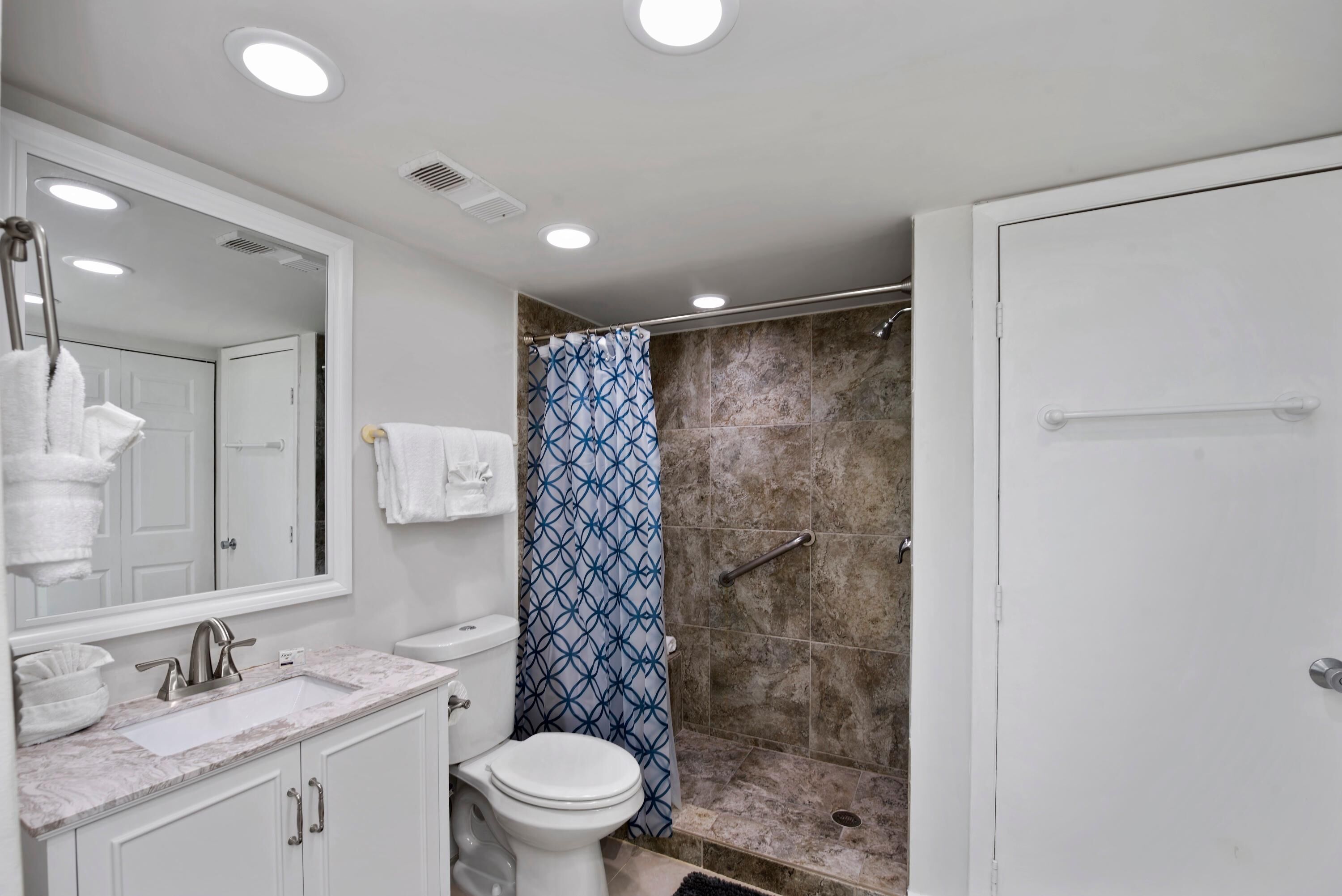 Un baño con lavamanos blanco, espejo y inodoro, ducha con cortina azul estampada, y puerta blanca.