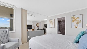A bedroom with a bed, a grey armchair, a view through a window, and a kitchen area in the background. - Estero Beach & Tennis 901-c (Fort Myers Beach)