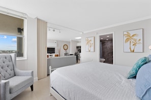 Quarto com cama, poltrona cinzenta, vista pela janela e área de cozinha ao fundo.