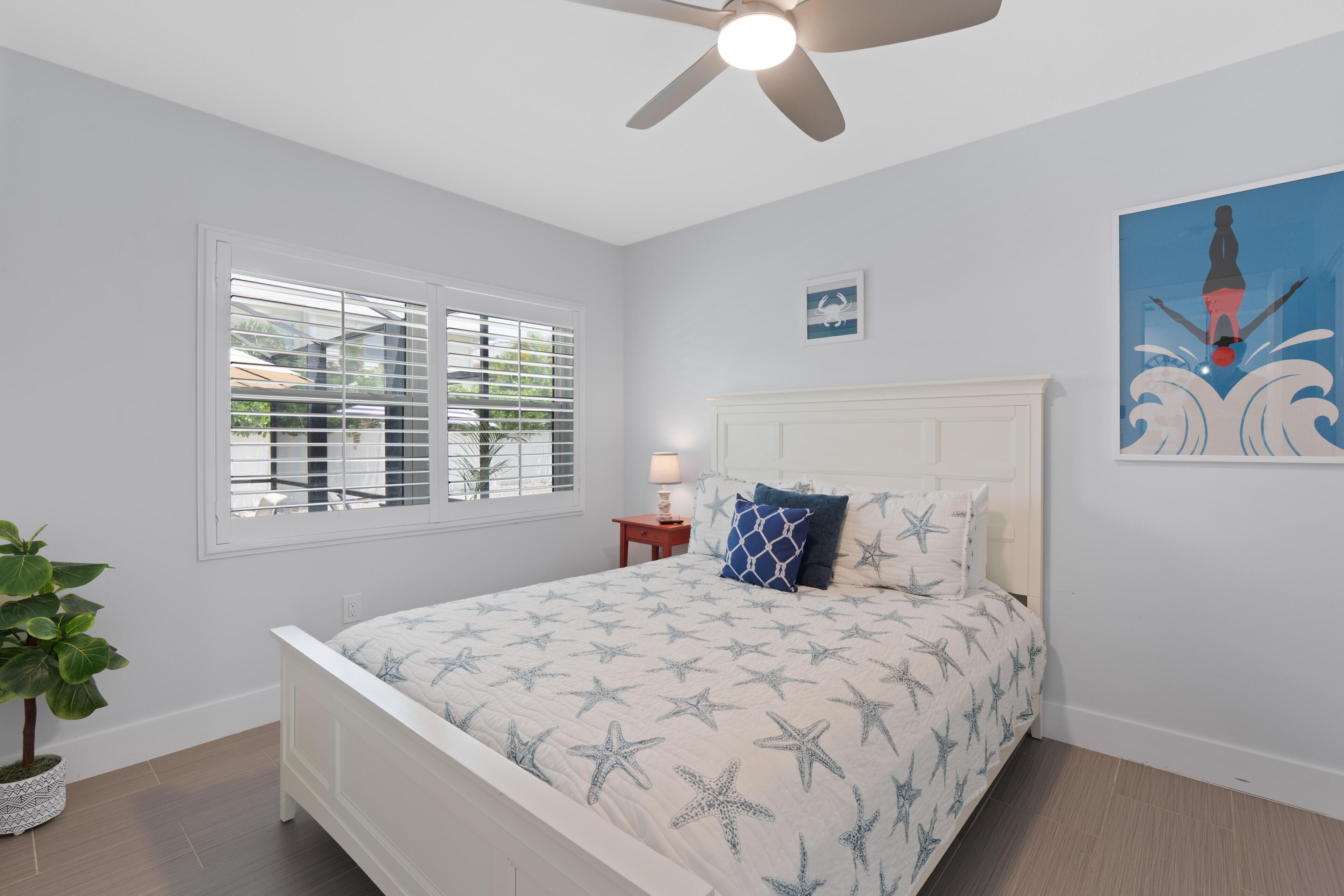 A bedroom with a bed, a ceiling fan, a window with blinds, a nightstand, a lamp, a wall-mounted artwork, and a potted plant.