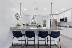 A modern kitchen with white cabinets, a central island with bar stools, stainless steel appliances, and pendant lighting. - Ocean Breeze (Fort Myers Beach)