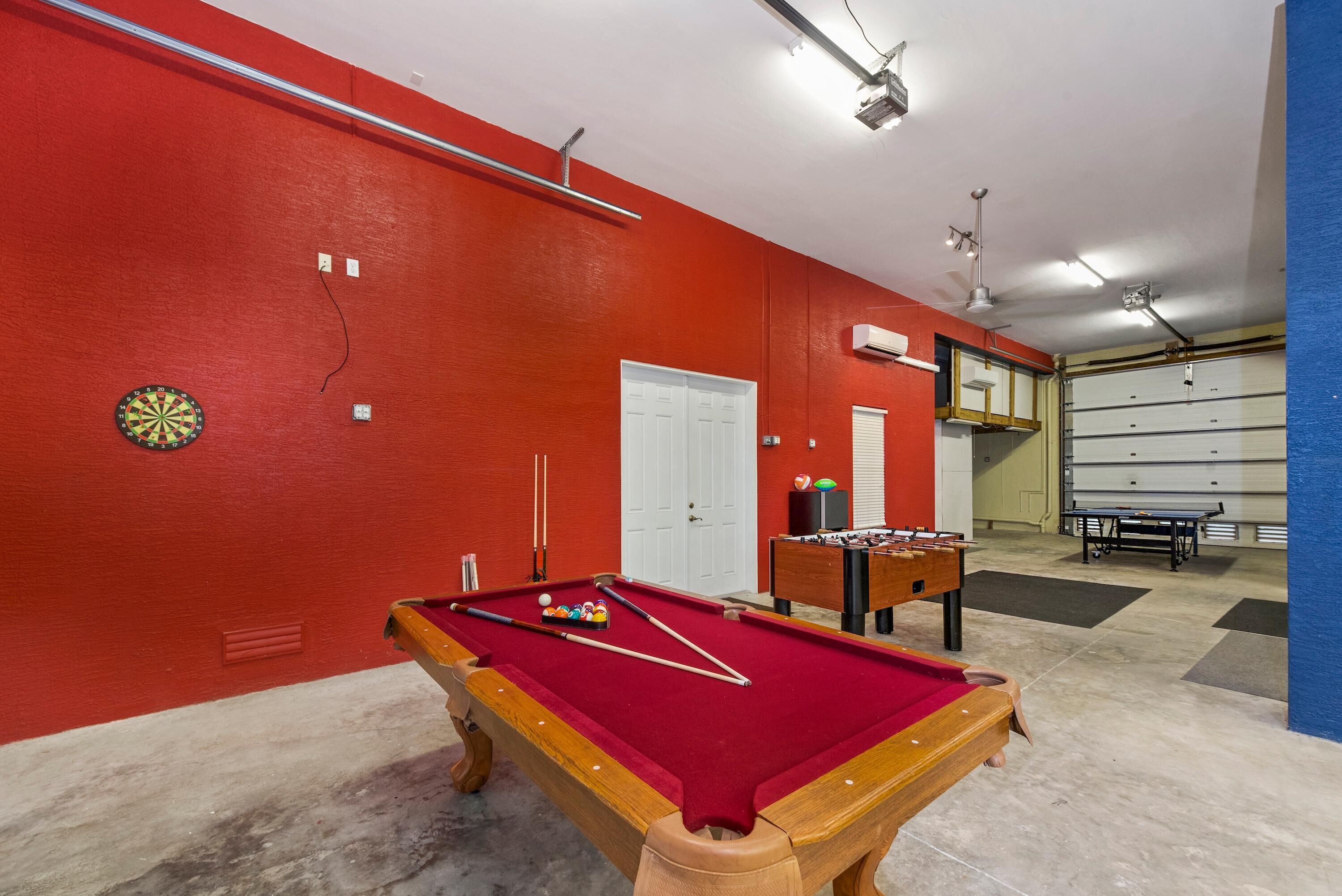 A pool table in a room with red walls, a dartboard, and a foosball table.