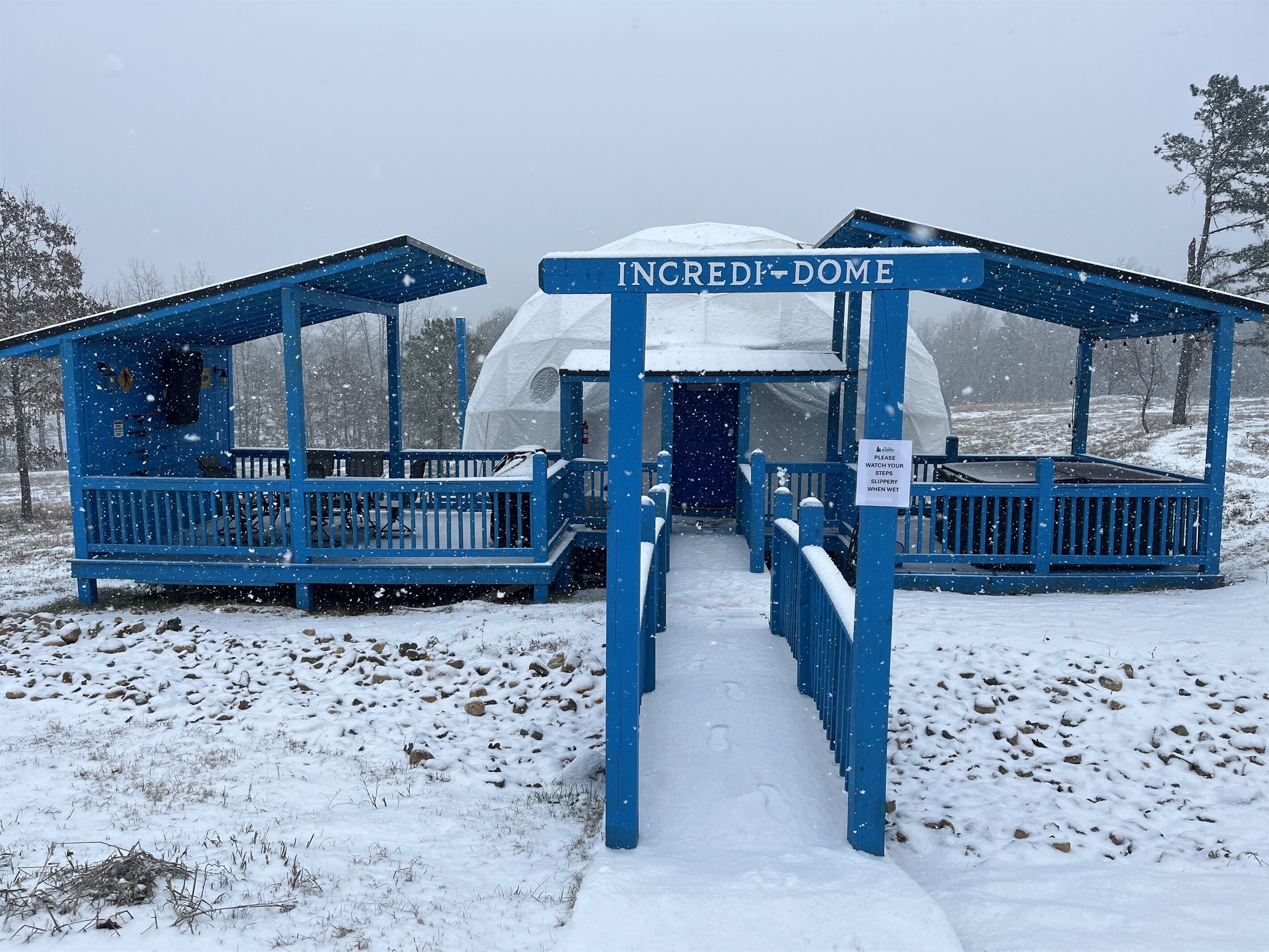 Entrada cubierta de nieve a una estructura en forma de cúpula con la leyenda “INCREDIBLE DOME”. Un letrero indica precaución al caminar sobre superficies mojadas.