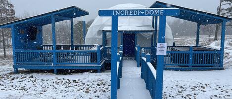 Entrada cubierta de nieve a una estructura en forma de cúpula con la leyenda “INCREDIBLE DOME”. Un letrero indica precaución al caminar sobre superficies mojadas.