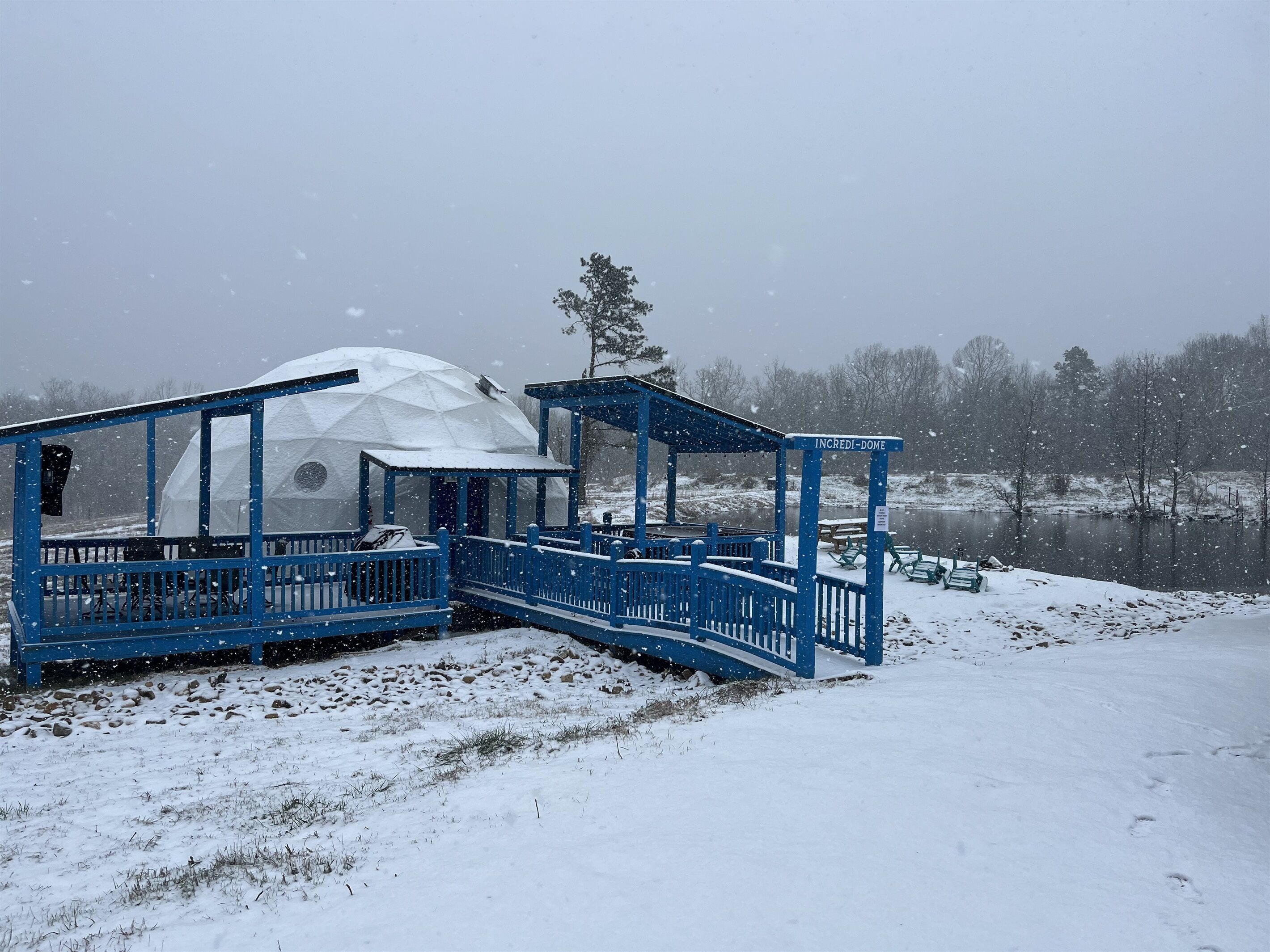 Una estructura abovedada con fachada azul y un porche cubierto, ubicada junto a un lago rodeado de nieve.