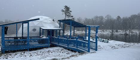 Una estructura abovedada con fachada azul y un porche cubierto, ubicada junto a un lago rodeado de nieve.