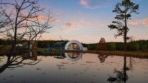 A dome-shaped building reflecting in a calm body of water, surrounded by trees and a clear sky.