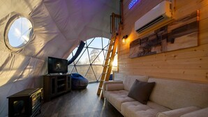 A cozy interior of a dome-shaped tent with a wooden interior, a sofa, a television, and a ladder.