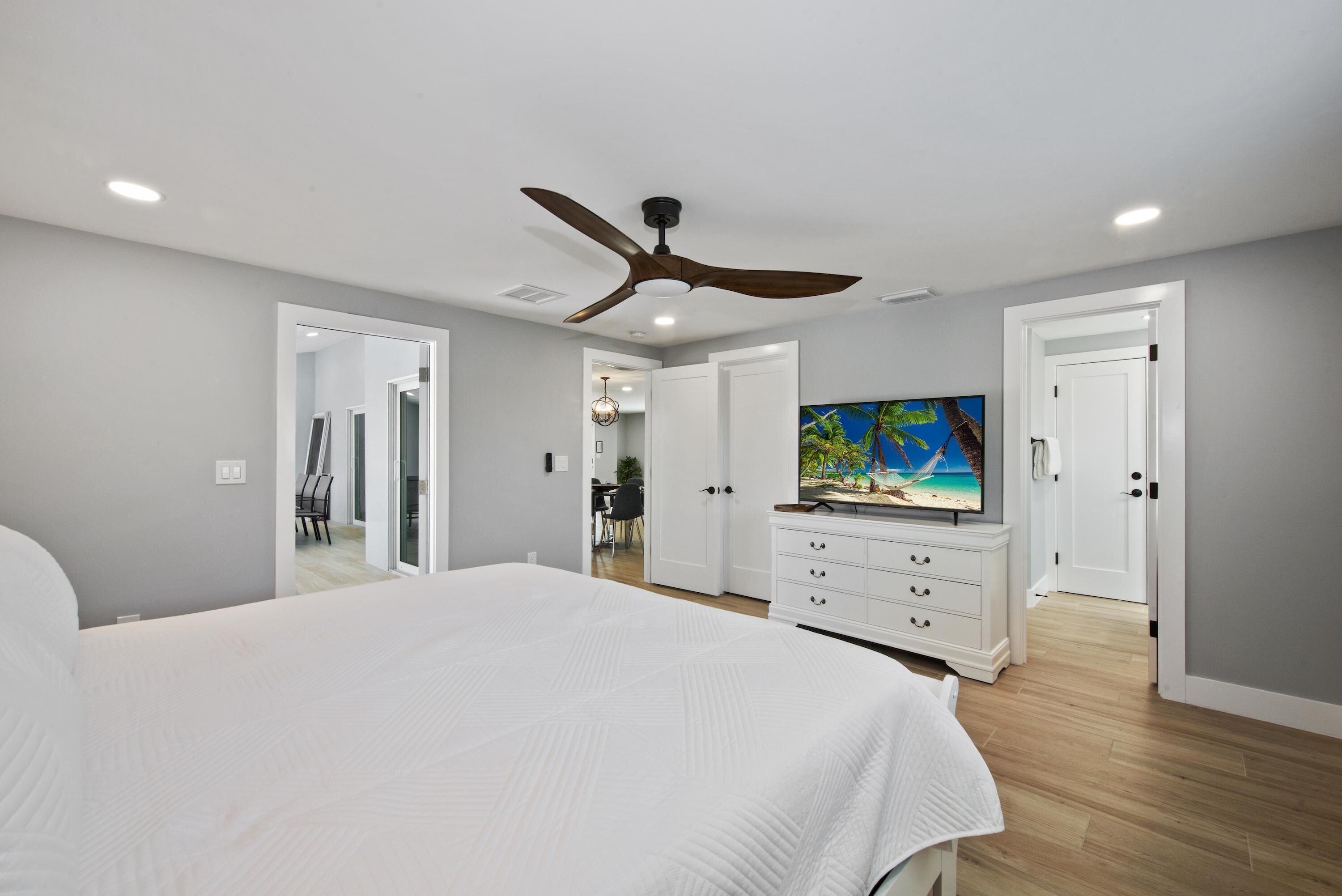 A bedroom with a large bed, a ceiling fan, a TV, and a painting of a beach scene.