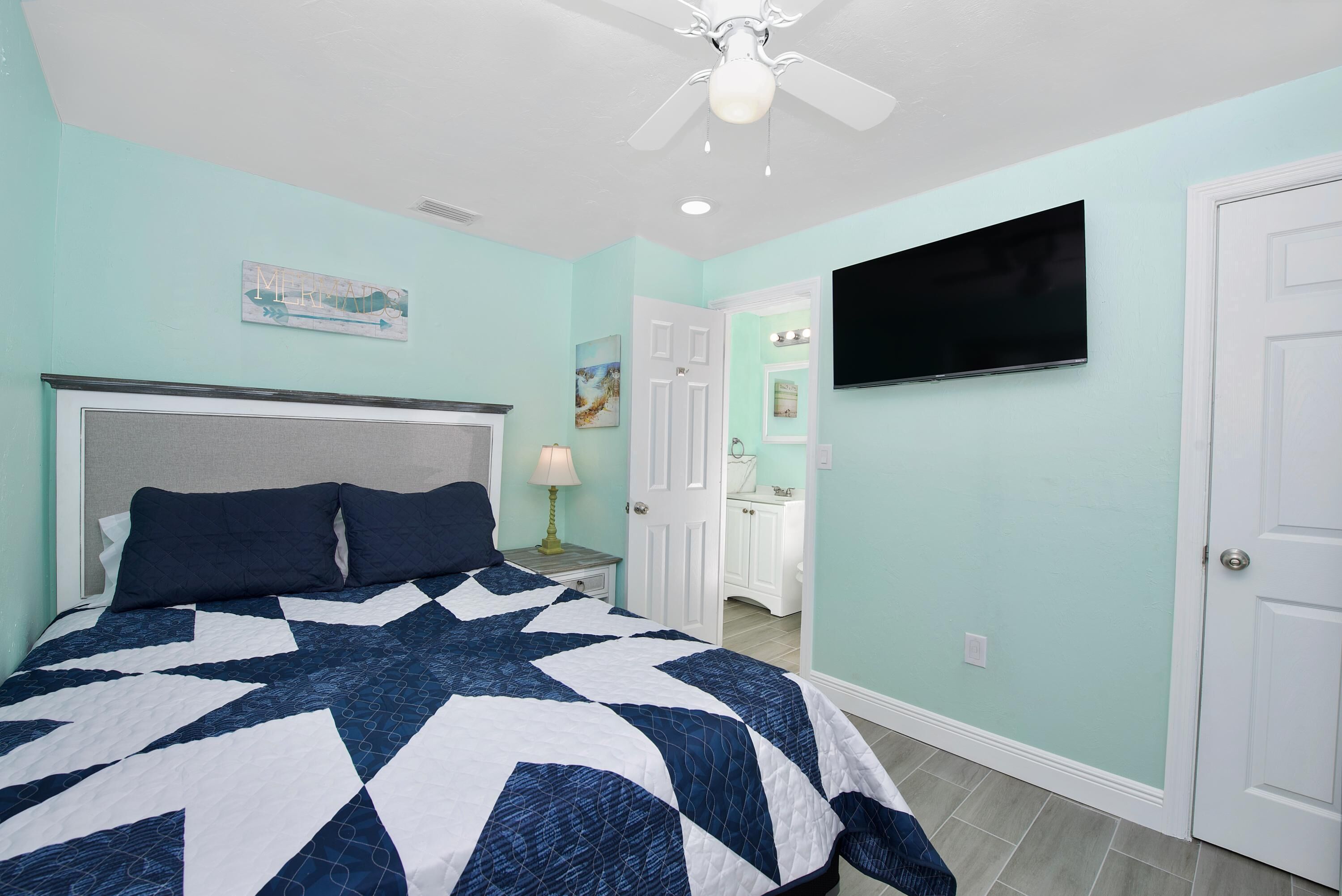 Un dormitorio con una cama grande, un ventilador de techo, un televisor montado en la pared y un baño visible a través de una puerta abierta.